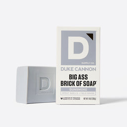 An gray brick of soap next to its packaging, which features the Duke Cannon brand logo and says "Gunsmoke. Spent Shells + Field Grass"
