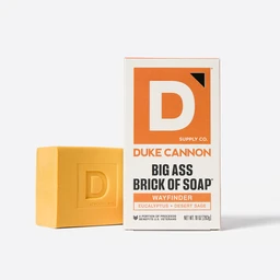 A yellow bar of soap next to its packaging, which is orange with the Duke Cannon logo and the product name 'Big Ass Brick of Soap - Wayfinder'.