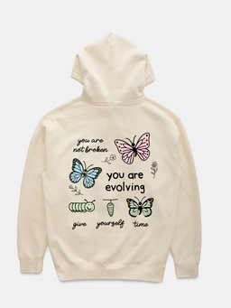This is a cream-colored hoodie with a relaxed, unisex fit. The back features a pastel butterfly graphic with the phrases “you are not broken,” “you are evolving,” and “give yourself time,” along with small botanical accents. Made from soft fleece-like fabric, it presents a casual, comfy style in light, neutral tones.
