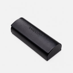 A compact, rectangular hard case with a rounded end cap and a curved lid. The visible material looks like smooth black leatherette with a subtle sheen, and the word “RANDOLPH” is embossed on the lid. The style is minimalist and unisex, suitable for a small accessory or eyewear; color is solid black.