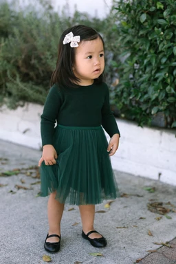 A young girl wearing a green knit dress with a tulle skirt, standing in a natural setting with trees and fallen leaves in the background.