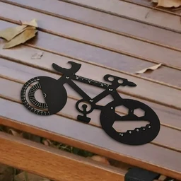 A black metal bicycle-shaped multi-tool with wrench openings, a protractor-style angle gauge and a sized cutout set, lying on a wooden bench with dry leaves.
