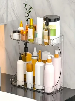 A two-tier transparent plastic countertop organizer holding white and amber cosmetic bottles, pump dispensers and jars on a dark surface against a light tiled wall.