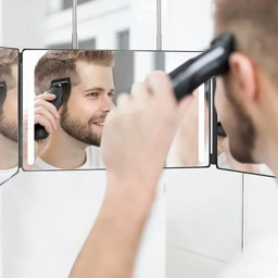 A man uses a black handheld clipper to trim his beard in a three-panel silver-framed mirror set on a white bathroom wall.