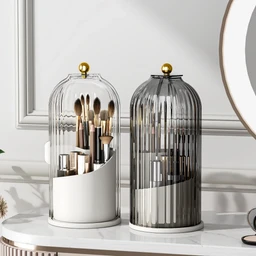 Two cylindrical domed organizers with gold knobs on a white marble vanity, one white with clear dome holding cutlery-like utensils and one grey-tinted clear ribbed cylinder, set beside a round mirror.