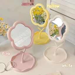 Three flower-shaped tabletop mirrors on slender curved stems and petal-scalloped bases in pink, yellow, and white plastic, set against a pastel background with fresh flowers.