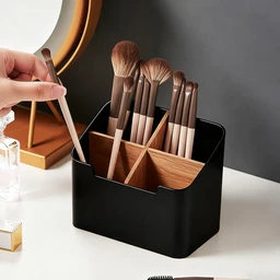 A black plastic desktop organizer holds wooden-handled makeup brushes in separate compartments on a white vanity beside a round mirror, while a hand reaches in to pull one brush out.