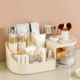 A cream-coloured plastic countertop cosmetics organizer with open compartments holding perfume bottles, skincare bottles, and makeup brushes, plus a clear front drawer, set on a pale surface against a beige wall.