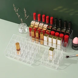 Three clear acrylic lipstick organizer racks with grid compartments holding gold-tubed red and black lipsticks on a pale green tabletop beside a small glass vase and makeup brush.