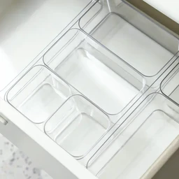 Several clear transparent plastic organizer bins of varying sizes sit inside a white drawer, showing a glossy acrylic-like finish.