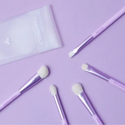 Five lilac-handled makeup brushes with white synthetic bristles lie on a lavender background beside a translucent white pouch printed with the TONYMOLY logo.