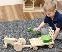 A young boy in a navy shirt plays on grey carpet with a pale wooden toy tractor, a wooden hay wagon stacked with green blocks, and more green blocks in his hand.