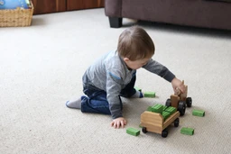 A toddler in a grey striped shirt and jeans plays on a beige carpet, pushing a natural-wood toy tractor and wagon loaded with green blocks, with more green blocks scattered nearby and a brown sofa in the background.