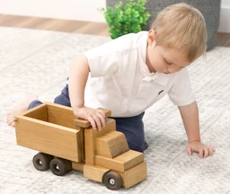 A young blond boy in a white polo shirt and navy shorts kneels on a cream rug beside a natural-finish wooden dump truck with dark wooden wheels and a tilted open bed.