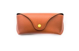 This is a hard-shell sunglasses case in a warm brown, pebble-grain leather-like material with an envelope-style flap and a small yellow snap button closure. The design is unisex, intended to protect eyewear, and the compact, curved shape suggests a snug fit for standard sunglasses. Notable attributes visible: textured brown exterior, curved flap, gold/yellow snap, and clean minimalist silhouette.