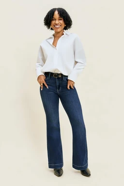 These are women's denim flare jeans in a petite length. The model pairs them with a white blouse and belt, showing a dark indigo, classic denim wash with a fitted mid-rise and a wide-flare leg that widens toward the bottom. The fabric appears to be sturdy cotton denim with subtle stitching and a clean, polished look.