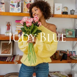 - Product: Judy Blue E-Gift Card (shown in a lifestyle shot). 
- Audience and styling: Appears targeted to women; a female model wears a cozy, casual look with a soft yellow fuzzy knit off-shoulder sweater and blue jeans, holding a bouquet of pink roses. 
- Notable attributes: Soft, chunky knit in pale yellow; casual denim; overall bright, feminine color palette and relaxed, everyday style.