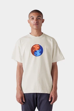 A person wearing the 686 Mens Bark and Bite Short Sleeve Tee by 686, featuring a cream color and a chest graphic of two stylized hands‚Äîone orange, one blue‚Äîreaching toward each other, stands against a plain white background.
