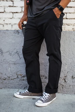 A person stands on a sidewalk wearing 686 Men's Everywhere® Pant - Relaxed Fit by 686, a dark shirt, gray Converse sneakers, a smartwatch, and holding a drink. The backdrop features a white brick wall and gray cement section.