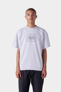 A person with short hair wears the 686 Men's Jib Flower Outline Short Sleeve Tee by 686— a loose, light-colored T-shirt with a black outline graphic on the chest—paired with dark pants, standing against a plain light background.