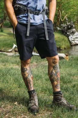 A person stands on grass with colorful leg tattoos, wearing 686 Men's Everywhere® Hybrid Short, hiking boots, a camouflage T-shirt, and a backpack. Trees and a fallen branch are visible in the background.