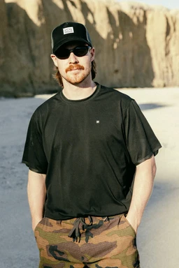 A bearded man in a black cap, sunglasses, and the 686 Men's Let's Go Tech Tee with camouflage pants stands on a sandy beach by tall cliffs, hands in pockets, looking at the camera‚Äîready for a spring-summer adventure.