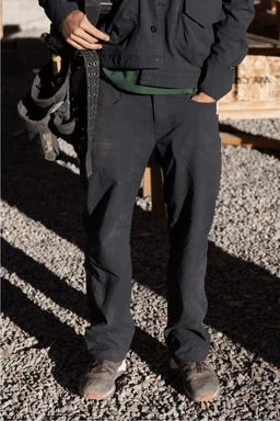A person stands outdoors on gravel wearing the 686 Men's Unwork® Pant - Relaxed Loose Fit, a work jacket, and sturdy shoes. With a tool belt at their waist and one hand in their pocket, their face is not visible.