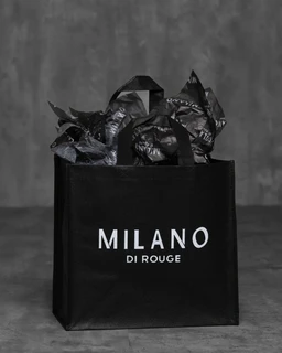 Black Milano Di Rouge reusable shopping tote made of synthetic non-woven polypropylene, with the Milano logo, filled with black tissue paper.