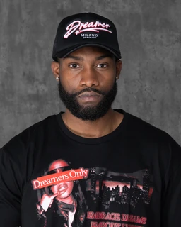 This is a black baseball cap (casual streetwear style) with a curved brim. The front features pink and white embroidery reading “Dreamer” with smaller text underneath, set against a black fabric. The model appears to be a man, suggesting the intended audience is men's; styling is casual with a graphic tee visible beneath.
