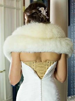 This photo shows a white satin corset viewed from the back, with beige cordage laces threaded through the eyelets and tied to shape the waist. The model appears to be an adult woman, styled in an elegant, formal look with a cream fur stole draped over her shoulders, suggesting a bridal or couture vibe. Visible attributes include the off-white fur wrap, the pale beige lace/cording for the corset, and the corset’s structured, voluminous cut with a back seam closure.